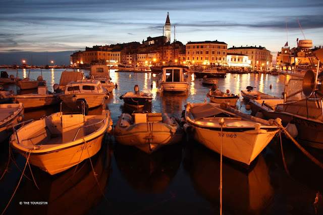 White fishing boats bobbing in a harbour during the blue hour, cottages, tavernas, and a hillside church dipped in the soft summer evening light.