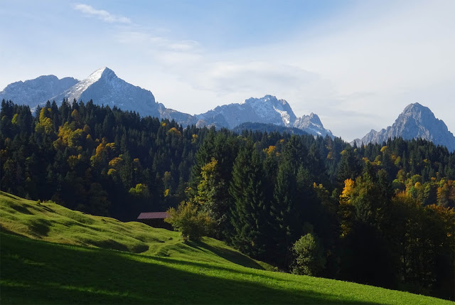 Wettersteingebirge mit schneebedeckten Bergspitzen, grüne Hügeln, braune Holzhütte und Wald 