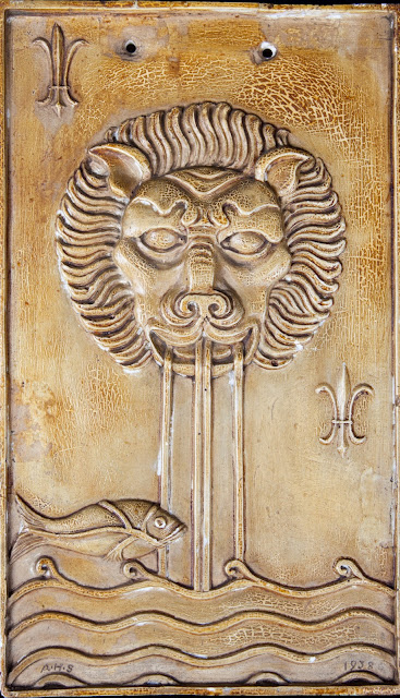 A rectangular plaque, with a lion's head and water pouring out into a body of water, and a fish swimming past