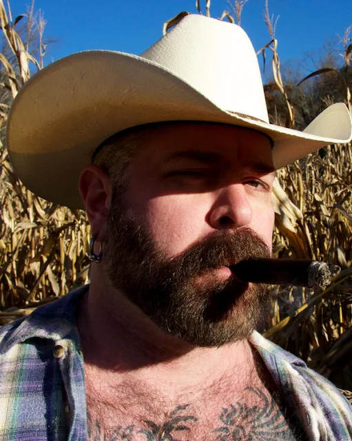 . Wearing a cowboy hat brown shirt and smoking a cigar in front of a cornfield