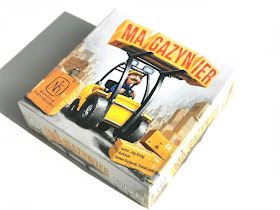 na zdjęciu pudełko gry Magazynier w kolorze białym z wizerunkiem uśmiechniętego magazyniera w kasku i z wąsem, jadącego wózkiem widłowym i wiozącego paletę ze skrzynkami, na których jest napis Magazynier