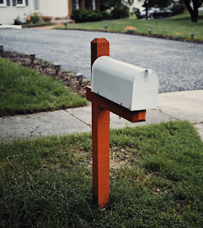 A white mailbox on a red wooden post next to a sidewalk and driveway.