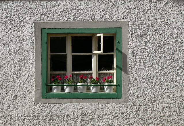 Janela com duas bordas, a primeira pintada de branca e a segunda de verde, há também vasos de flores decorando a faixada, está em uma parede branca.