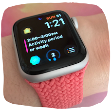 Photo of an Apple Watch. The strap is pink braided. On the digital watch face is the time, date, a calendar event and app symbols.