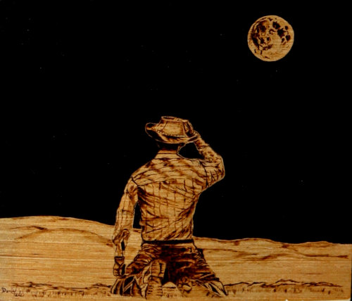 Painted woodburning of a cowboy gazing at the moon in a night sky.