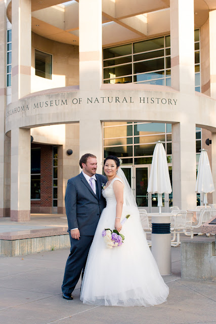 He has his arm around his bride, while gazing lovingly at her.  She stands facing him with her head turned towards the viewer.  They are standing in front of the Sam Noble Oklahoma Museum of Natural Histoy, where their reception is being held.  They have become husband and wife today and their connection and excitement is palpable.  Taken on a day in early September with the sun low in the sky.