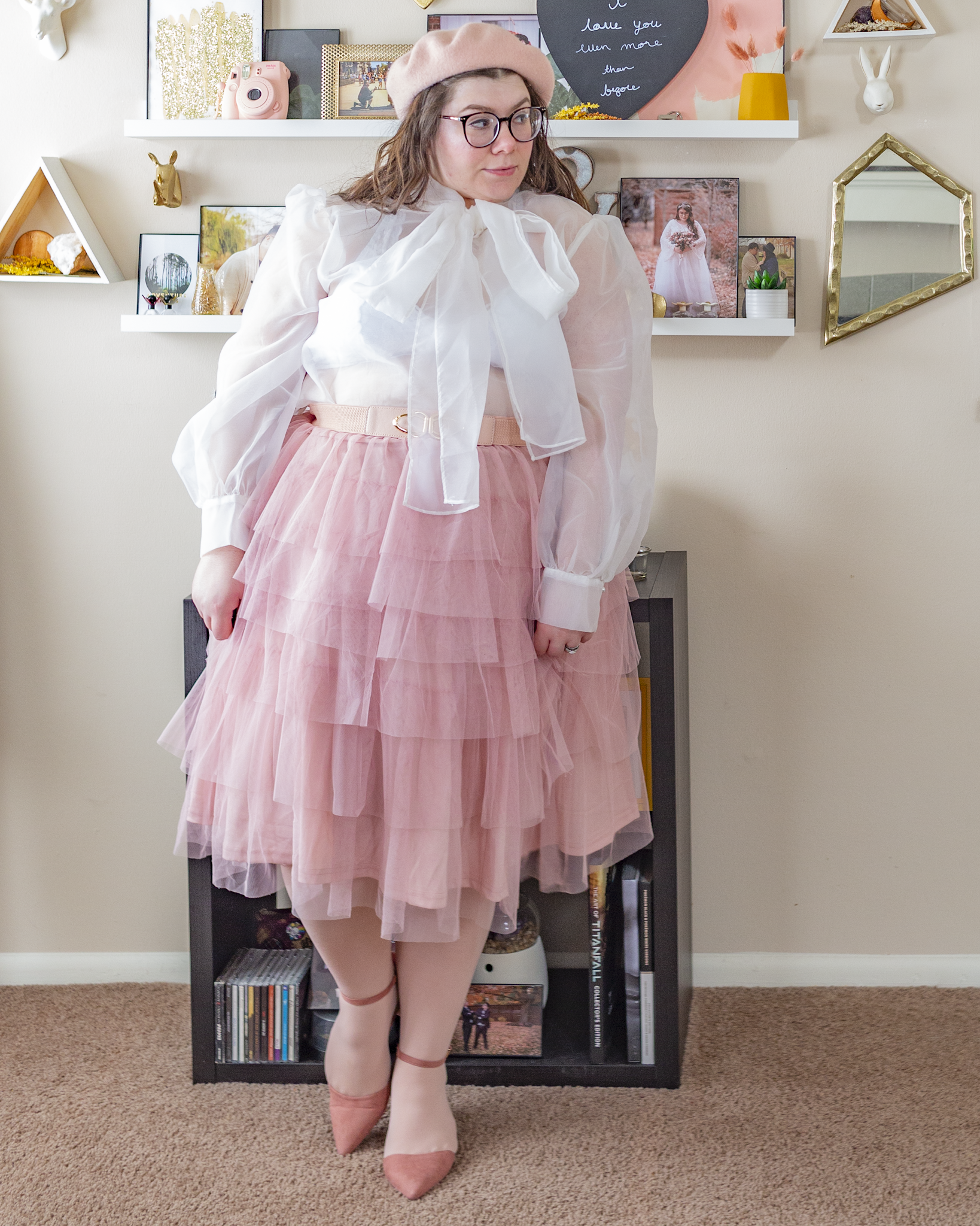 An outfit consisting of a pastel pink beret, a white sheer long sleeve billow sleeve blouse with a connected tie neck, tied into a bow, tucked into a dusty pink tiered tulle midi skirt with pastel pink tights and pink ankle strap pointed toe heels.