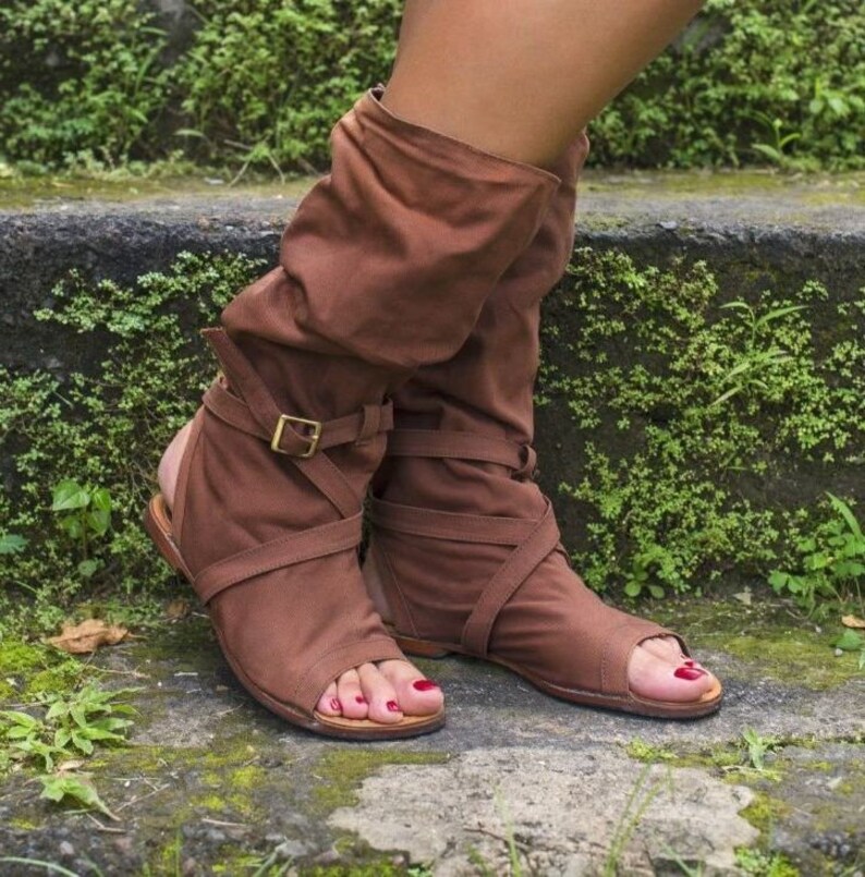 Open toe and heel boots, brown, in suede leather. They are adjusted with cross straps on the instep and at the ankle. The wearer has red nail polish and is posing with one foot slightly raised.