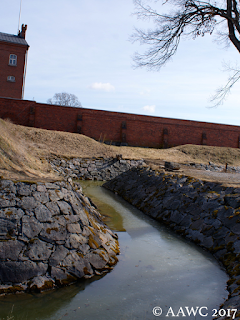 A side-on view of Hämeen linna, including part off the moat in the foreground and small of the red brick-like castle in the background.