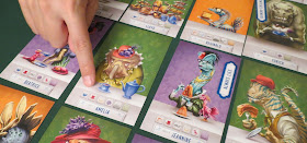 A close-up of some of the cards laid out in a grid. A few examples that can be seen include Amelia, a leptoceratopsian dinosaur wearing an light olive green dress and a red hat with purple flowers, looking somewhat grumpy as she sits at a table with a kettle of tea and a teacup; and Jeannine, a blue dilophosaurus in a forest green outfit wearing a small red hat adorned with black feathers, lots of gold necklaces glasses, and lipstick, holding a large gelatin, with a small feathered pink dinosaur on the table beside her. Someone is pointing to one of the icons on Amelia's card, and a token that says 'Always Lies' has been placed on Jeannine's card.