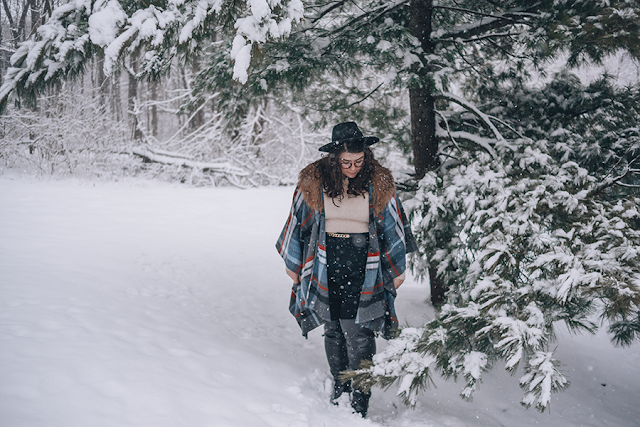 An outfit of a black wide brim hat, gray, red, and blue plaid shawl with brown faux fur collar, cream sweater, black button up mini skirt, and black over the knee boots in snow.