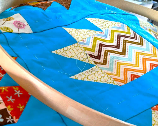 A section of quilt with a blue background, a threaded needle stuck in it and part of a quilting hoop shown on top.