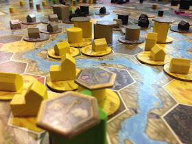 A game of Terra Mystica in progress. The board, made up of a series of hexagonal spaces, each representing a different terrain type, some with discs to indicate that it has been changed to a different terrain type, is covered with wooden pieces of various types, some of which have cardboard tokens indicating that they've been turned into cities.