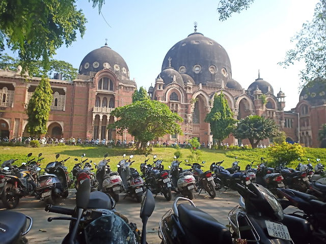 Red sandstone and black domed Arts Faculty building at MS University Baroda, with garden in front and two rows of parked motorcycles