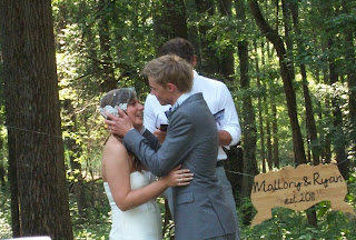 At the outdoor alter, the groom is about to kiss the bride on the forehead. The pastor looks on.