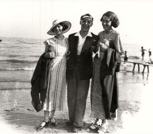 Two women and a man linked arms walking along the beachfront, they are smartly dressed