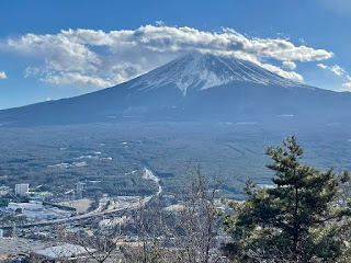 A landscape photo of snow-capped Mount Fuji, surrounded by clouds, with roads and countryside in the foreground.