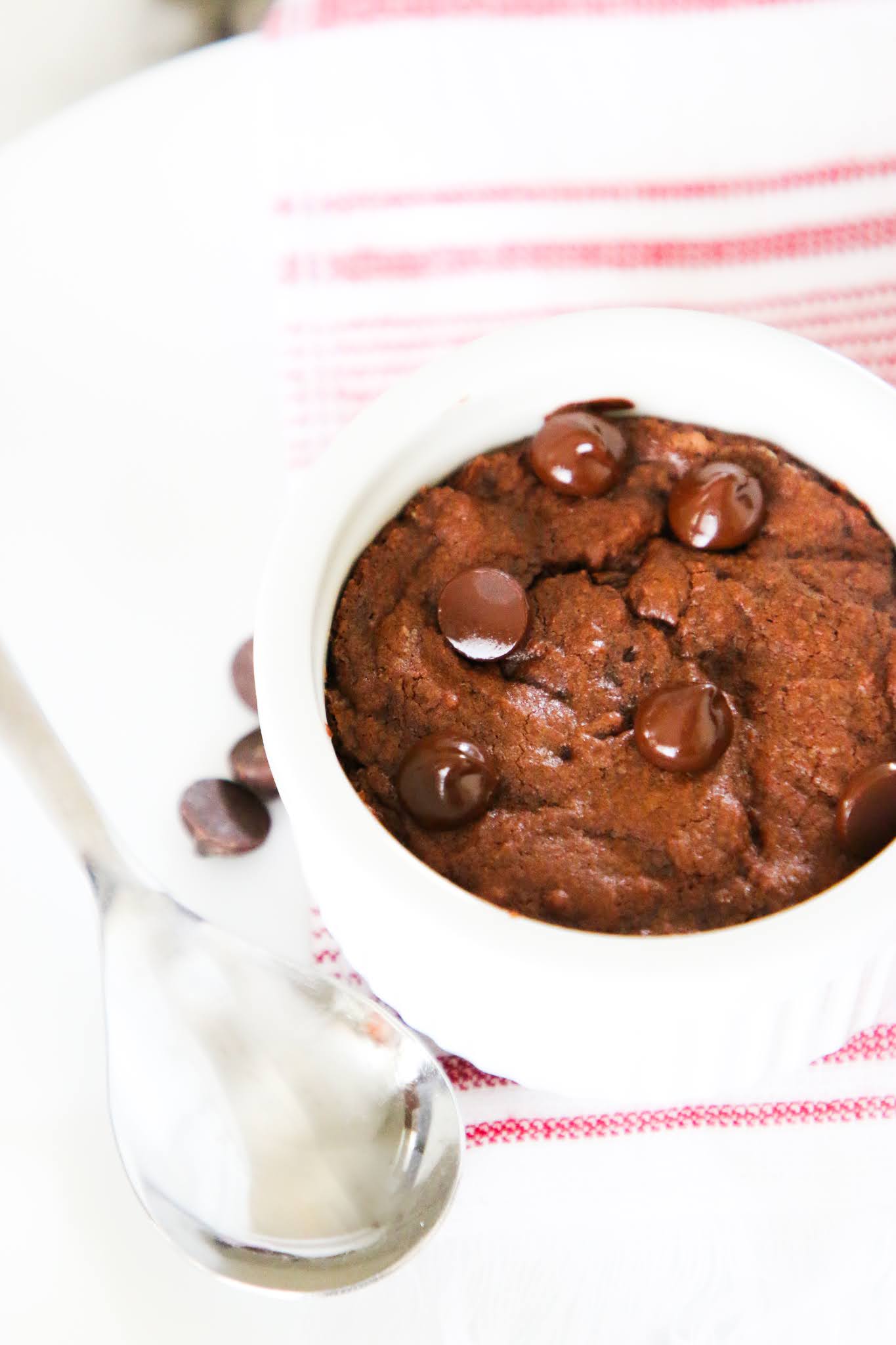 Brownie baked in a small white ramekin on top of a red dish towel placed on a white plate with scattered chocolate chips on a white marble table.