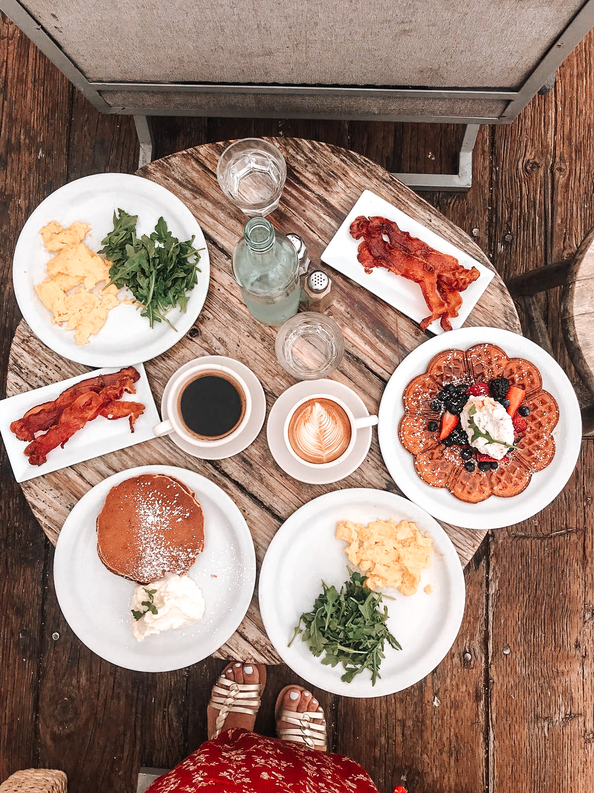 table en bois au restaurant malibu farm à los angeles avec assiettes blanches dans lesquelles sont disposés des gaufres et des fruits rouges, des pancakes des oeufs brouillés du bacon et de la salade roquette