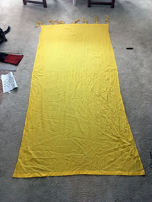 A bright yellow, narrow, rectangular gauze curtain with fabric ties at the top, laid flat on tan carpet.