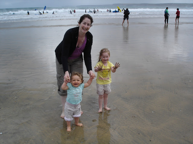 Brown haired mum on sand as white sea rolls in behind, holding hands of one happy one year old girl and three year old daughter stands next to her showing off big smile and sandy hands