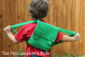 The boy is wearing a red shirt.  He is stretching his arms out, along with the green straps of his backpack.