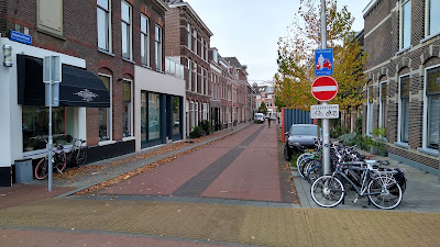 A narrow street with houses both sides. The red road has a wide grey stripe in the middle. Cycle parking is to the right. There is a no entry except cycles sign.