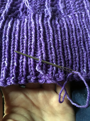 A close-up of the edge of a section of purple knitted ribbing, with a silver yarn needle threaded with the free end of yarn and inserted under one stitch, about an inche from the edge..