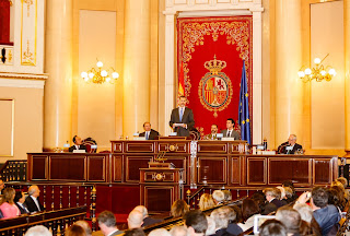 Rei Filipe VI discursando no Senado da Espanha em 2015. Ao fundo, vê-se um repostero com as armas dessa instituição, que são as mesmas do estado num escudo oval, debruado de azul, rodeado pelo colar da Ordem do Tosão de Ouro e, acima, por um listel de azul que sai da coroa, carregado da legenda SENADO DE ESPAÑA em letras de ouro. Imagem disponível no site institucional.