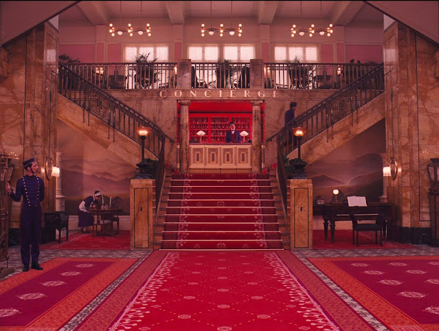 Cena de "O Grande Hotel Budapeste". Mostra o lobby do hotel. As paredes são de madeira e o chão está completamente coberto com tapete vermelho. Há uma escada no meio do salão que se divide em duas. É um ambiente luxuoso.