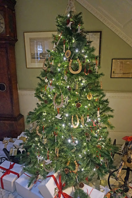 Christmas Tree decorated with lights in a red and white theme including decorations of dogs, deer, horse shoes and birds, with presents wrapped in white & red in a room with light green painted walls, a grandfather clock on the left and a old bicycle at at Stourhead in Wiltshire