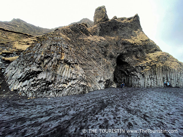 A grey rainy sky over a cave surrounded by tall narrow basalt columns on a black lava beach.
