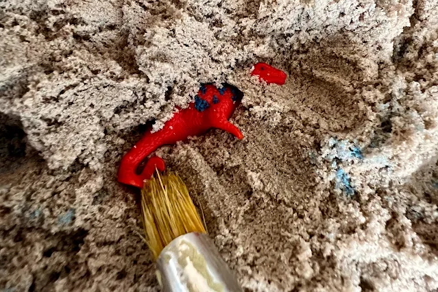 A close up of a paint brush being used to uncover a toy dinosaur in sand