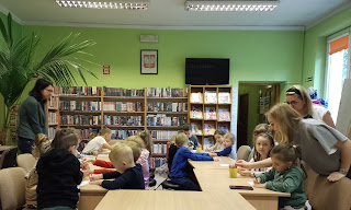 tło: regały z książkami. Pani bibliotekarka i panie przedszkolanki wśród przedszkolaków siedzących przy stolikach.