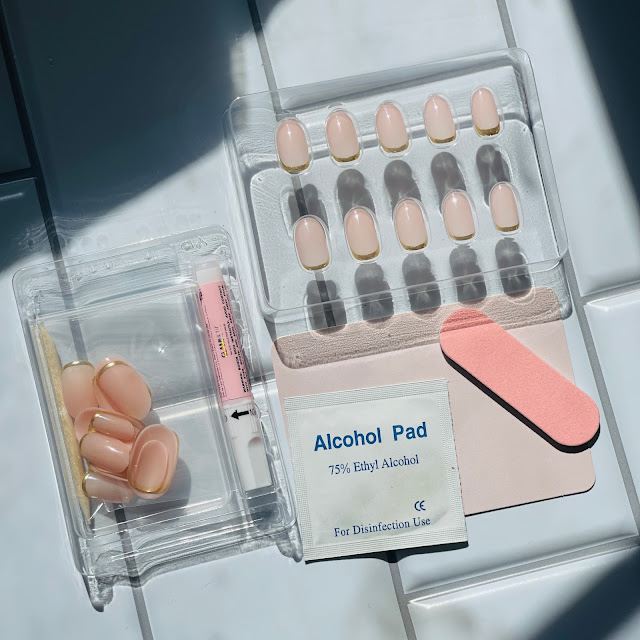 Contents of a box of glamnetic nails on a white tile background including package of nails, tube of glue, alcohol wipe, and file