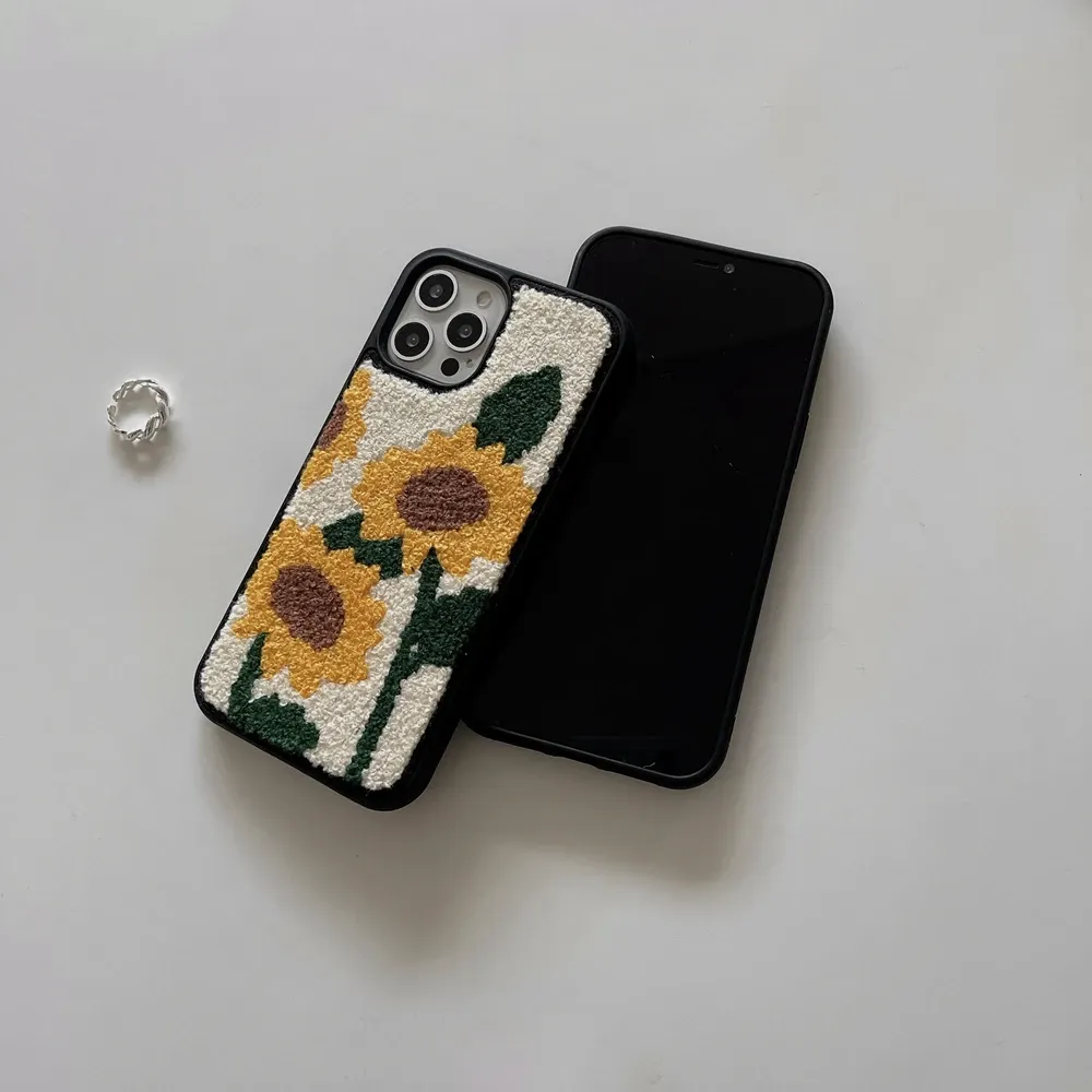 A white iPhone case with a colorful embroidered sunflower design on the back. The sunflower has a yellow center with orange and brown accents, and green leaves. The case appears to be handcrafted and has a slightly textured surface.