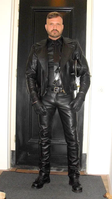 Handsome man standing in the hallway wearing full black leather gear Muir cap under arm