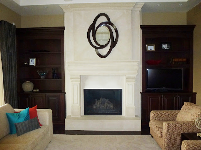 close up of COCOZY reader's great room with almost empty built in bookshelf, fireplace with molded mantel, neutral sofa with three accent pillows one blue, red and patterned and two arm chairs