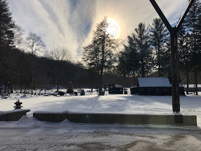 Snow-covered landscape at Drake Well Museum & Park with cloud-muted sun casting shadows of bare trees and evergreens. The replica derrick and oil lease office are also visible with snow on their roofs.