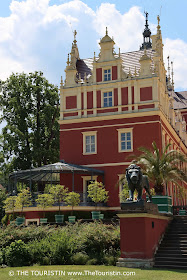 The sculpture of a lion on a pillar of a large staircase in front of a reddish painted renaissance castle.