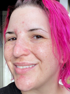 Lara, a pale skinned person with neon pink chin length hair, smiling at the camera after applying moisturiser. Her head is turned slightly and her skin is glowing.