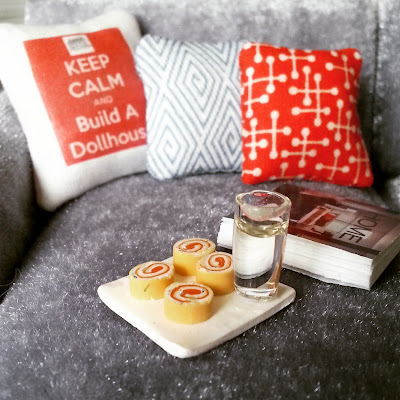 Corner of a one-twelfth scale modern miniature sofa with several cushions, a drink in a glass, several pieces of cake and a book.