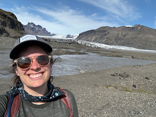 Mel, brunette with baseball cap and sunglasses taking a selfie in front of a glacier on a sunny day