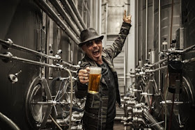 Rudolf Schenker dentro de uma fabrica de cerveja segurando um caneco de cerveja com uma mão, fazendo pose de grito e o simbolo rock and roll com a outra mão. 