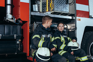 Photo of two firefighters in uniform sitting on a fire truck and talking.