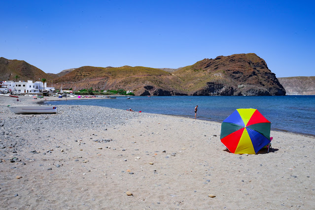 Playa con una sombrilla de colores sobre la arena y las azules aguas del mar y los acantilados rocosos de fondo