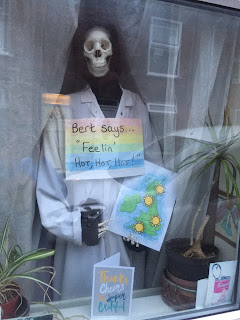 A skeleton wearing a white lab coat and holding a sign 'Bert says...Feeling' hot, hot hot!'