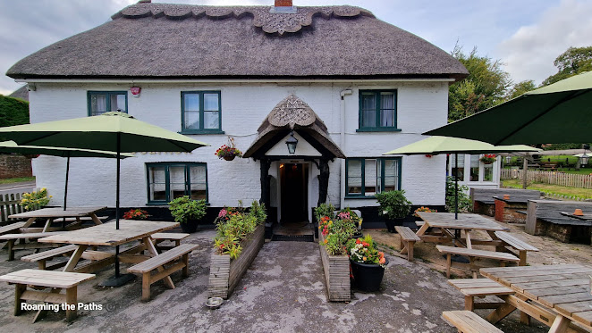 Front Entrance with an old thatched porch, the fromnt of the pub havinga  thatched roof too. A patio garden with large benches adn parasols, with containers and baskets full of colourful and fragrent flowers