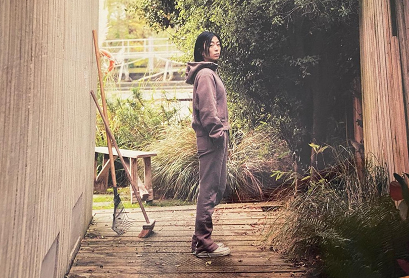 A shot of Hikaru Utada in a back garden, stood with their hands in their pockets. Hikaru Utada is wearing the same brown sweat set that they are wearing on the Bad Mode album cover.