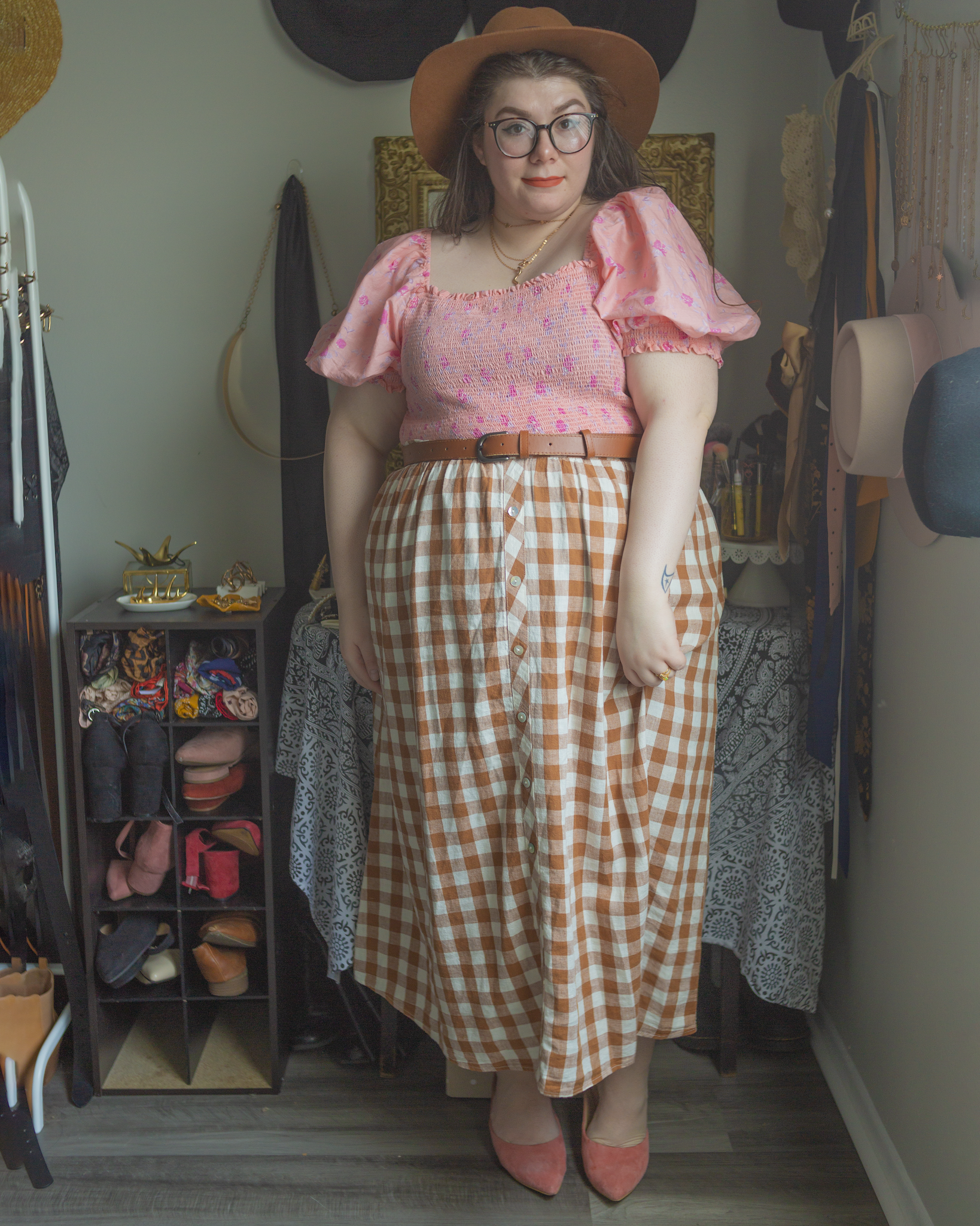 An outfit consisting of a brown panama hat, a pastel floral pink shirred crop top with puffy cap sleeves under a brown gingham button down midi skirt and pastel pink heeled clogs.